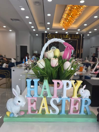 The image displays an Easter-themed decoration on a white counter within the "Center Nail Bar" in Naples, United States. The primary subject is a white woven basket containing artificial white and light pink tulip-like flowers, green foliage, small white blossoms, and two speckled artificial eggs (one yellow, one white), with a pink bow tied to its handle. In front of the basket, colorful block letters spell "HAPPY EASTER" on a light green base. A white ceramic rabbit figurine is positioned on the left with three miniature pastel eggs (yellow, blue, pink) at its feet, and a smaller white ceramic rabbit is on the right.
The background shows the interior of the nail salon, identifiable by a sign reading "CENTER NAIL BAR." Multiple workstations with dark blue chairs and gold accents are visible. Several individuals, consistent with patrons and staff, are present and appear to be receiving or providing nail services. The ceiling is white with recessed lighting and a large, ornate, gold-toned chandelier.