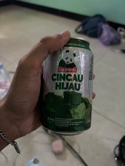 A person's hand holds a can of "Cincau Hijau" soda. The can is silver with green text and a panda graphic. The background is blurred but appears to be an indoor setting with a tiled floor and a dark green wall. The can is covered in condensation. There is a water bottle visible on the left side of the image. The image does not contain enough information to determine the location in Meteseh, Indonesia.