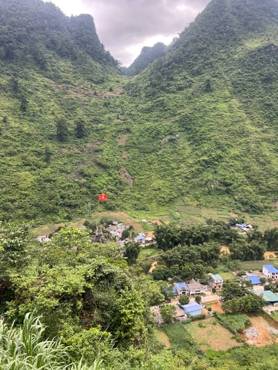 A high-angle shot captures a verdant mountain landscape with a small village nestled in the valley. The mountains are densely covered in lush green foliage, with some rocky outcrops visible. A red flag with a yellow star, the flag of Vietnam, flutters from a pole in the middle ground, positioned above a cluster of houses. The village itself consists of a collection of homes with brightly colored roofs, surrounded by agricultural fields. Bamboo trees and other vegetation line the edges of the village and fields. A muddy river or stream flows through the lower right portion of the frame. The sky is overcast with clouds.