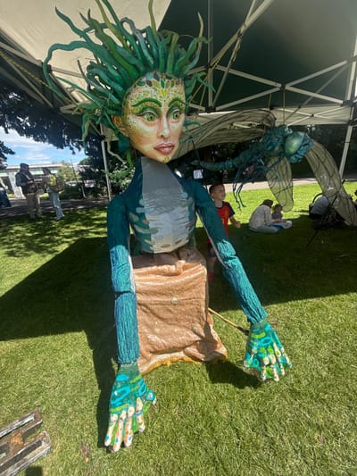 In Leominster, United Kingdom, a large, artistic puppet resembling Medusa, adorned with green, vine-like hair, is displayed outdoors on a sunny day. The puppet has a detailed face with green and yellow markings, large eyes, and pale skin. It wears a teal, scale-patterned body and a tan, textured skirt. To the right of the Medusa puppet, a large, teal dragonfly puppet is suspended from a green tent structure. In the background, several people are visible, including a young boy in a red and blue shirt and two women sitting on the grass with a child. The scene is set on a grassy field beneath a tent, with trees and buildings visible in the distance.