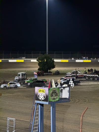 The image depicts a night scene at the Dirt Oval Route 66 in Joliet, Illinois, United States of America. The foreground shows a section of a metal fence and a ladder leading up to a platform with signs that read "DART OVAL ROUTE 66" and "MONSTER TRUCK MAYHEM". Several vehicles are visible on a dirt track in the background, including a white tow truck with people on it, a green pickup truck, a black SUV, and a white car. A few people are standing near the vehicles and the fence. The track is surrounded by a fence, and in the far distance, a crowd of spectators can be seen standing along a tiered area. A tall light pole with a bright light illuminates the scene.