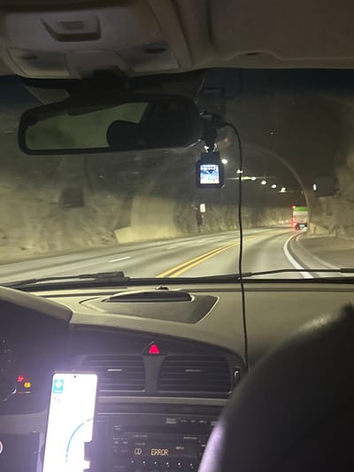 An interior view from the driver's seat of a vehicle traveling through a tunnel. The tunnel walls are rough and textured, with lights spaced along the ceiling. The road ahead is paved with double yellow lines in the center and a white line on the right side. A dashcam is mounted on the windshield above the rearview mirror, displaying a live feed. On the dashboard, a smartphone is mounted, showing a navigation app. The car's interior includes an airbag indicator light, a hazard light button, and a car radio with an "ERROR" message displayed. The overall lighting suggests it is nighttime or the tunnel is dimly lit. The image does not contain any information about the city of Stavanger, Norway.
