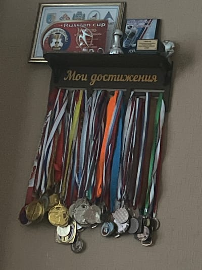 This image shows a collection of medals and awards displayed on a wall. The medals have various colored ribbons and metallic charms. Some of the medals are attached to a dark wooden plaque with gold lettering. The plaque has the Russian text "Мои достижения" which translates to "My Achievements". To the left of the plaque, there is a framed certificate or award, which appears to be for the "Russian cup" and has some emblem on it. There are also some other awards and trophies visible. The scene is indoors, with a plain, textured wall as the background. The lighting suggests it is daytime. The overall impression is one of accomplishment and recognition.