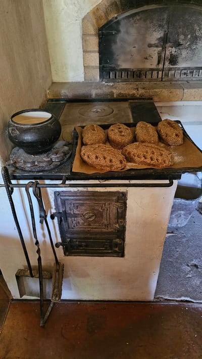 In the city of Sodeliškiai, Lithuania, an indoor scene depicts a rustic, traditional oven. The oven, likely an ancient appliance, is constructed from white plaster and features a dark metal door with intricate detailing. To the left of the oven door, a set of wrought iron fireplace tools, including a poker, tongs, and a brush, are neatly hung. Above the oven, a dark metal stovetop is visible, with a black cast-iron pot positioned on a round burner. Adjacent to the pot, on a black baking tray lined with parchment paper, sit seven loaves of dark, rustic bread, each with a slightly oval shape and visible grains. The background shows the arch of the oven's firebox, framed by stone and plastered walls. The floor in the foreground is a reddish-brown material, possibly wood or aged metal, with patches of grey concrete to the right. The overall atmosphere is one of historical domesticity and home baking.