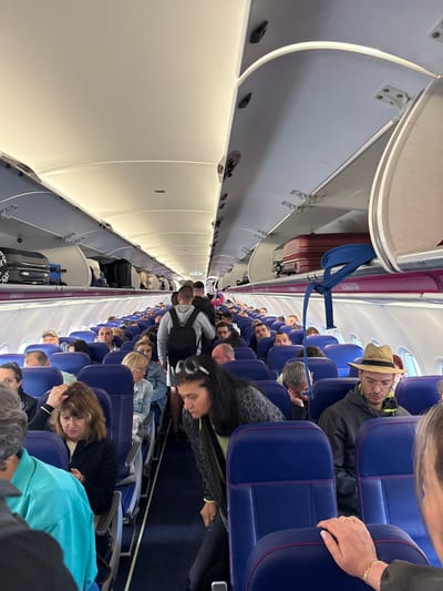 This image shows the interior of an airplane cabin, with rows of blue seats occupied by passengers. Overhead luggage bins are open, containing suitcases and bags. The aisle is partially visible, with some passengers standing and others seated. The lighting appears to be the standard interior lighting of the aircraft. There is no indication of the city of Künersberg, Germany, or any specific location within it.