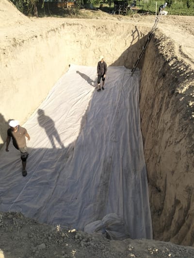 In Aktam (Tashminvody), Uzbekistan, a deep, rectangular excavation is visible, its sandy walls showing signs of erosion. Inside the pit, a large, white geotextile fabric is spread across the base. Two men are present in the excavation. One man, wearing a brown polo shirt and shorts, stands on the geotextile, facing towards the left. Another man, wearing a patterned shirt and shorts, is positioned further up the slope on the right side of the excavation, also on the geotextile. A wooden ladder leans against the right wall of the excavation, leading up to the ground level. The surrounding environment appears to be rural, with vegetation and some wooden structures visible on the distant upper edge of the excavation. The scene suggests ongoing construction or excavation work.