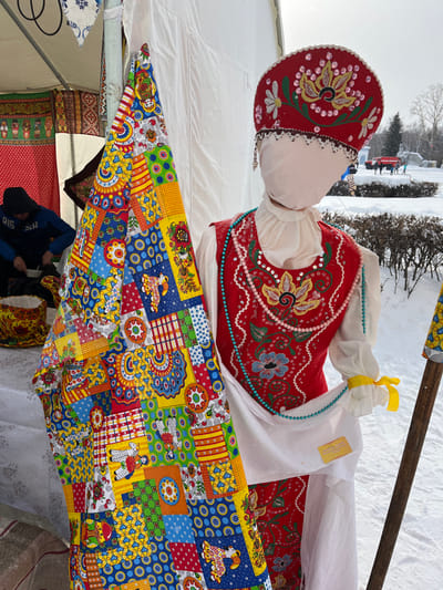 The image shows a large, anthropomorphic doll or effigy, prominently displayed outdoors in what appears to be a winter setting. The doll is oriented vertically, with its head at the top and the long, flowing skirt extending downwards.
The doll's head is a simple white sphere, topped with a striking red traditional Russian headpiece, possibly a kokoshnik. This headpiece is intricately embroidered with floral patterns in white, yellow, and green, and adorned with numerous small, shiny elements that resemble pearls or beads, particularly around the edges.
Its upper body is clad in a white long-sleeved blouse with puffy shoulders and ruffled cuffs, one of which has a yellow ribbon tied around it. Over the blouse, the doll wears a vibrant red vest or sarafan top, also richly decorated with embroidery. The embroidery on the vest features green leafy motifs, blue and yellow flowers, and scattered silver sequins or beads. Two strands of beads are draped around the doll's neck: an inner strand of light pink beads and an outer strand of brighter blue beads. A small, rectangular yellow tag is visible on the white fabric of the doll's right sleeve.
The lower half of the doll is dominated by an expansive, colorful skirt made of a patchwork-style fabric. This fabric is composed of numerous small squares and rectangles, each featuring different patterns. These patterns include geometric shapes (circles, squares, stripes), stylized floral designs, and various pictorial elements such as birds, roosters, sheep or lambs, horses with riders, and what appear to be traditional Russian matryoshka dolls. The colors are very bright and varied, including blues, yellows, reds, greens, and oranges.
The doll is positioned under a white tent or canopy, parts of which are visible in the upper left and lower left of the frame. Behind the doll, a rough wooden pole or beam runs diagonally upwards and to the right. The background outside the tent reveals a snowy landscape with snow-covered ground and bare trees. In the distance, faintly visible, are some structures and possibly people or vehicles, suggesting a public event or gathering. The lighting suggests it is daytime, possibly overcast given the soft light and lack of harsh shadows.
In the lower left corner, beneath the doll's skirt, other patterned fabrics are visible, possibly indicating additional merchandise or decorations within the tent. Blurred figures, likely people, can also be seen in the lower left section, partially obscured by the fabrics.
The overall appearance of the doll, with its traditional Russian attire and the snowy setting, strongly suggests it is an effigy, possibly for a celebration like Maslenitsa (Shrovetide), which marks the end of winter in Slavic cultures.