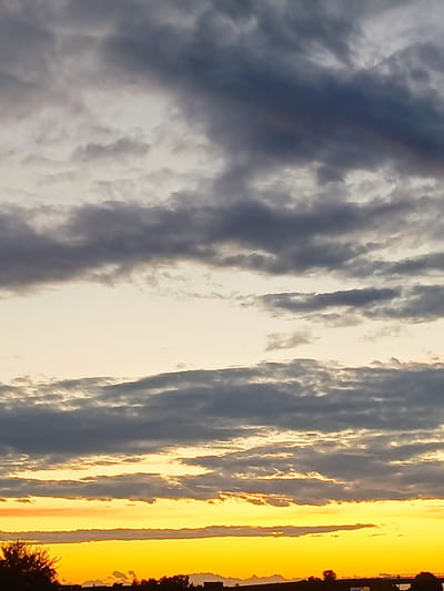 The image depicts a sunset or sunrise over a rural landscape, likely in the vicinity of Somaglia, Italy. The sky is filled with a dramatic display of clouds, with layers of dark gray and lighter gray formations stretching across the frame. Below the cloud cover, a vibrant band of golden-yellow light illuminates the horizon, indicating the position of the sun. The lower portion of the image shows a silhouetted treeline and a dark, indistinct landscape that could include buildings or infrastructure. The overall impression is one of a natural, atmospheric event.