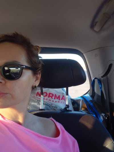 A woman with her hair pulled up and wearing sunglasses is in the passenger seat of a car. She is wearing a pink top. In the background, there is a headrest and a shopping bag with the word "NORMA" visible on it, suggesting it is from a supermarket. Part of another bag with blue trim is also visible. The scene appears to be from the interior of a vehicle, with a reflection of the road visible in the woman's sunglasses.