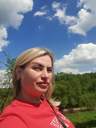 A woman with fair skin and blonde hair is taking a selfie outdoors. She is wearing a red shirt. The background includes a blue sky with white clouds and a dense green forest. The location is likely near a wooded area, given the vegetation. The image is rotated slightly, with the sky appearing to the left and the trees to the upper right. Without further information, I cannot confirm that this image was taken in the city of Avdot’ino, Russia.