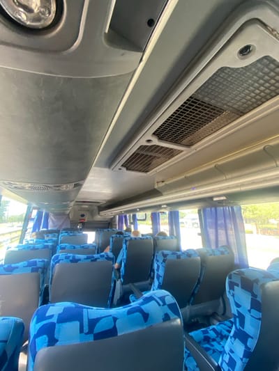 The image displays the interior of a passenger bus in Belén de Escobar, Argentina, captured from a forward position looking aft along the central aisle. Multiple rows of passenger seats are visible, characterized by a blue and light-blue geometric patterned fabric on the cushions and upper backs, contrasted with dark grey lower backs and bases. Approximately three to five passengers are discernable, seated from behind in the mid-to-rear sections of the bus. One individual in the mid-section wears a blue shirt and has light-colored hair. The ceiling is grey, featuring two rectangular air conditioning vents with grilles and white overhead compartments. A partial view of a circular overhead light fixture is present in the top-left. Blue curtains are partially drawn over the windows on the right side. Through the windows, daylight is evident, revealing a roadside barrier on the left, and green trees alongside a light-colored vehicle in the background on the right.