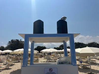 The image depicts a beach scene in Costa Rei, Italy, under a clear blue sky with a few distant clouds. In the foreground, a white and blue wooden structure stands, topped with two large black speakers. One of the speakers has a circular, white and black device mounted on it. The structure is situated on a sandy beach dotted with numerous empty white and beige beach umbrellas and loungers, suggesting a currently unoccupied area. In the background, a line of green trees borders the beach. A sign with a red circle and a diagonal line over a swimming figure, along with Italian text, is affixed to the front of the wooden structure.