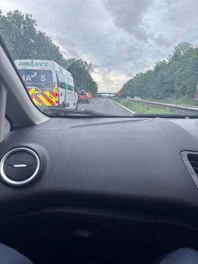 From inside a vehicle, a rainy highway scene is visible. An ambulance is in the adjacent lane, with its lights visible. Other vehicles are further ahead. The sky is cloudy, with trees lining the side of the road. The vehicle's dashboard, including an air vent, is prominently displayed in the foreground. The image appears to be taken from the driver's seat.