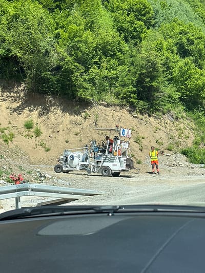 The image shows a construction scene on a steep, wooded mountainside in Crkvine, Montenegro. A white, multi-wheeled vehicle with equipment, possibly for road maintenance or construction, is parked on a gravel surface. Two individuals are present: one wearing a bright orange and yellow vest and pants stands near the vehicle, while another person, partially visible, is seated in the vehicle. The environment consists of a dirt embankment covered in lush green trees and vegetation, with scattered rocks and debris on the ground. A metal barrier is visible in the foreground. The overall impression is of ongoing work in a natural, mountainous setting.