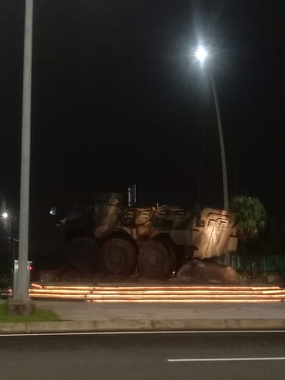 The image displays a stationary armored personnel carrier (APC) at night in Kosambi Barat, Indonesia. The multi-wheeled military vehicle, appearing to be a permanent display or monument, is positioned on an elevated, rocky base. Its body is camouflaged in dark, muted tones. Six large wheels are visible on the side facing the viewer. The scene is illuminated by artificial light sources: a prominent streetlight with a distinct starburst effect is visible in the upper right, and another less bright light appears in the upper left. Horizontal, parallel glowing lines, emanating from ground-level linear lights, are present in front of the vehicle's base. In the immediate foreground, a paved road with a white lane marking is visible, separated by a curb and a sidewalk from the display area. A tall, light-colored pole, likely a streetlight support, extends vertically along the left side of the frame from the bottom. Green foliage is discernible behind and to the right of the armored vehicle. No individuals are present in the scene.