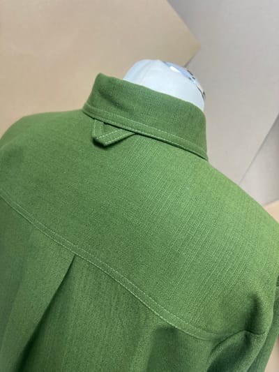 A close-up shot focuses on the collar and shoulder area of a green, textured garment, possibly a shirt or jacket, displayed on a partial mannequin form with a white head. The fabric appears to be a woven material. White stitching is visible along the seams. The background consists of a beige surface, likely a table or display platform, and a blurred white wall. The image does not depict a location in the city of Kirklees, United Kingdom, or contain any information to infer that location.