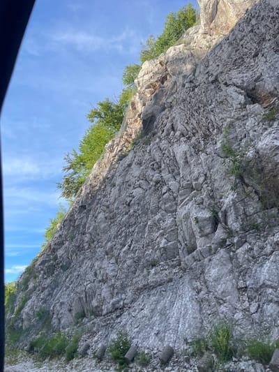 The image shows a steep, rocky cliff face with a protective wire mesh system installed. Green trees and vegetation grow at the top and along the base of the cliff. The sky is blue with some scattered clouds. The scene appears to be along a road, as suggested by the protective barriers at the bottom of the frame and the overall perspective which seems to be taken from a vehicle. The location is identified as Obzovica, Montenegro.