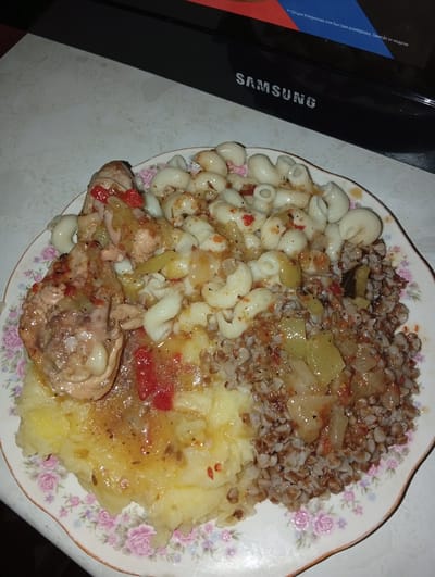 A close-up shot shows a plate of food, likely a meal. The plate is white with a delicate floral pattern around the rim, featuring pink roses and smaller purple flowers. The food is arranged in distinct sections. On the left side of the plate, a piece of cooked chicken, possibly a thigh or drumstick, is covered in a glistening sauce with visible pieces of red pepper and other vegetables. Adjacent to the chicken is a mound of creamy, yellow mashed potatoes, also with some sauce and seasoning on top. In the center and right of the plate, there are two other food items. One appears to be elbow macaroni, cooked and mixed with a sauce containing similar red pepper and vegetable pieces as the chicken. The other is a generous portion of buckwheat, a dark grain, also mixed with some vegetables and sauce. The overall presentation suggests a hearty, home-cooked meal. The lighting in the image is dim, indicating it was likely taken indoors at night or in a poorly lit room. There is no visible indication of the weather. In the background, a black Samsung electronic device is partially visible, with a hint of a screen displaying some text. Some of the visible text in Russian appears to be related to product features. The scene is set on a table with a textured, light-colored surface. There are no people visible in the image.