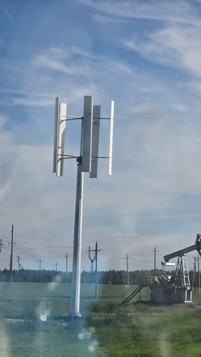 In an open field under a cloudy blue sky, a vertical axis wind turbine stands tall. The turbine features three rectangular blades arranged vertically on a central pole, with supporting horizontal rods. To the right, a pumpjack, characteristic of an oil extraction site, is visible. The landscape is dotted with power lines and poles, indicating a rural or industrial setting. A dense line of trees forms the horizon in the background. The foreground is a green field with some areas of dry grass and a few circular light flares.