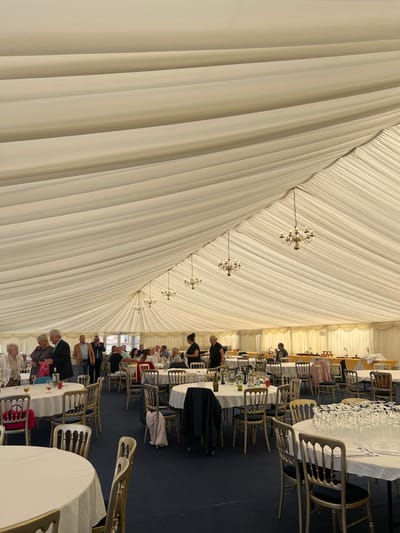 The image depicts the interior of a large event marquee, situated in the city of Scarborough, United Kingdom. The marquee's ceiling and walls are lined with a pleated, off-white fabric. The floor is covered with a dark blue carpet. Multiple brass-toned chandeliers, each fitted with several candelabra-style bulbs, are suspended from the ceiling at regular intervals.
The space is furnished with numerous round tables, each covered with a white tablecloth. Surrounding these tables are light-colored chairs, possibly gold or silver, featuring distinctive scalloped or fluted backrests. Several tables contain bottles of wine and water, along with various glasses. A table in the mid-right foreground holds a large quantity of empty wine glasses stacked neatly, indicating preparation for service.
Approximately 25-30 adults are visible within the marquee, dressed in smart casual to semi-formal attire. People are observed standing in small groups, conversing, particularly on the left side of the frame, and also seated at various tables throughout the venue. Two individuals in black uniforms, consistent with event staff, are visible near the center-right, engaged in what appears to be attendance to the tables. In the background, long rectangular tables, partially covered with yellow and white tablecloths, suggest a buffet or serving area. The scene portrays an active social or private function within a temporary event structure.