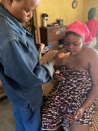 In Panyam, Nigeria, an individual wearing a denim jacket is applying makeup to another person who is wrapped in a patterned cloth and wearing a pink headscarf. The makeup application appears to be focused on the lips. The environment seems to be an indoor setting, possibly a home, with furniture and a clock visible in the background. Another person in a red shirt and green headscarf is partially visible in the background.