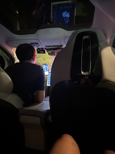 A person is seated in the passenger seat of a car. The person is wearing a dark-colored shirt, and is looking towards a dashboard screen. A screen is also located to their left. The interior of the car is primarily shades of gray and black. The location of the image is in Yuhuatai District, China.
