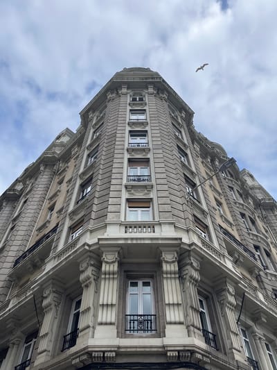 A low-angle shot captures a tall, ornate building in La Coruña, Spain. The building features a light-colored facade with repetitive window patterns and intricate architectural detailing. A bird is visible in the upper left of the frame against a cloudy sky. The building appears to be a historic structure.