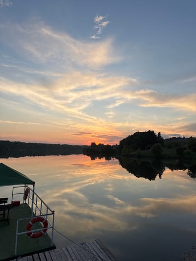 The image depicts a serene sunset over a calm lake, as seen from a dock in the city of Kamysh, Russia. The sky is a canvas of soft oranges, pinks, and blues, with wispy clouds stretched across it. The sun, a bright orange orb, is setting on the horizon, casting a warm glow over the scene. The lake perfectly mirrors the sky and the lush green trees that line the opposite shore.
In the foreground, a green-canopied structure with white railings is visible, suggesting a seating area or a small boat dock. Two orange life preservers are hanging on the railings, and a red emergency box is placed on a bench. Part of a wooden dock is also visible, leading away from the structure. The overall atmosphere is one of tranquility and natural beauty, typical of a Russian lakeside during the evening hours.