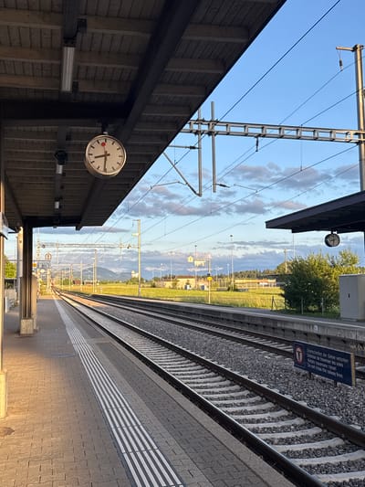 This image, correctly oriented, captures a serene late afternoon or early evening scene at a train station platform in Fehraltorf, Switzerland. The platform, paved with interlocking tiles and featuring a white striped safety line, occupies the right side of the frame, bathed in warm, golden sunlight. Two sets of railway tracks, complete with sleepers and gravel ballast, run parallel to the platform.
On the left, a station canopy with a wooden slatted wall is in partial shadow, contributing to the sense of time passing. A prominent classic Swiss railway station clock, characterized by its red second hand, is mounted beneath the canopy, indicating the time is around 4:50 PM or 5:00 PM. No people are present, emphasizing the quietness of the station.
Above the tracks, an extensive overhead catenary system of metal poles and wires is visible against a clear blue sky with scattered, wispy clouds. In the distance, beyond the tracks, the landscape features green fields, scattered buildings, and a backdrop of distant hills or mountains.
A notable detail is a blue warning sign positioned next to the tracks, which displays text in multiple languages: "Überschreiten der Gleise verboten," "Défense de traverser les voies," "Vietato traversare i binari," and "Do not cross the railway lines," along with a pictogram of a crossed-out person, clearly prohibiting track crossing. A smaller yellow sign marked "B" can also be seen further down the tracks, likely denoting a platform section. The overall mood is peaceful and calm under a pleasant sky.