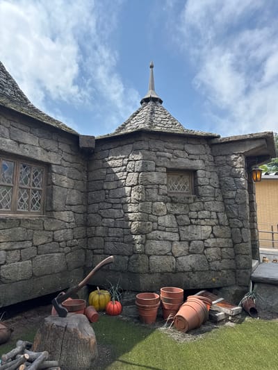 The image shows a stone building with a pointed roof, likely a themed architectural structure, under a partly cloudy sky. The building facade consists of irregularly shaped stones fitted together, and there are two windows with leaded glass. The exterior decor features stacked terracotta pots, pumpkins, an axe leaning against a tree stump, and other rustic items. The scene suggests an outdoor display or set-up, possibly a fantasy or village theme. The location in Nerima, Japan, cannot be verified from the image alone.