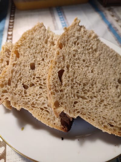 Three slices of artisan bread are arranged on a white plate with a gold rim. The bread has a light brown crust and a porous, airy interior. The slices are slightly overlapping, with the largest slice in the foreground on the right. In the background, a patterned tablecloth is visible. There is a small green speck on the plate to the left of the bread. The image appears to be taken indoors. The location of Денисово, Russia is not discernable from the visual information provided in the image.