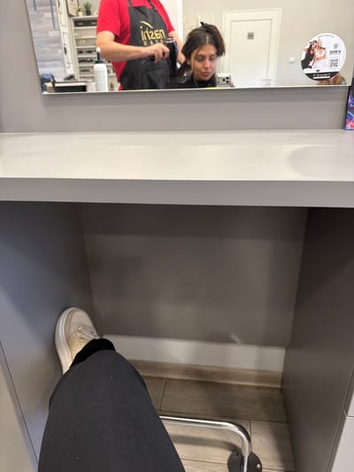The image depicts an interior scene, identified as a hair salon in Karnalovo, Bulgaria, captured from a first-person, seated perspective. In the immediate foreground, a person's right leg, clad in black trousers and a light beige sneaker, is visible, extending into a grey cabinet recess. A light grey countertop spans the mid-section of the image. Reflected in the mirror above this countertop, a male individual wearing a red shirt and a black apron with a gold "iritzen hair" logo is observed operating a black hairdryer, directed towards the head of a female client. The client, whose dark hair is partially secured with a white clip, is looking downwards, holding a mobile phone with a silver case in her left hand. A white squeeze bottle is visible on the counter in the reflection. Background elements reflected in the mirror include a white shelving unit containing various products, a white door with a small square sign, and a circular sticker adhered to the mirror. This sticker features images of two women, text including "www.f-expert.bg", "BLOOM 16", "F Expert", and a QR code. A fragment of a brightly patterned purple object is visible on the far right edge of the mirror reflection. The visible flooring beneath the cabinet is light wood laminate, and a curved chrome footrest bar is partially visible under the countertop.
