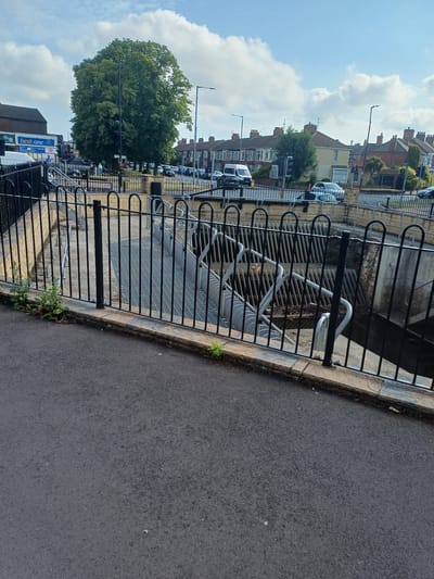 This image captures a view of the River Tees outfall structure, located near the A66 in Middlesbrough, United Kingdom. In the foreground, a paved walkway borders a black metal fence that encloses the outfall. The outfall itself is characterized by a grated metal walkway leading down into a channel. This channel contains a series of curved metal baffles, designed to regulate water flow. The background reveals a street scene with parked cars and residential buildings typical of the area. A large, leafy tree stands prominently, adding a natural element to the urban environment. The sky above is partly cloudy, suggesting fair weather.