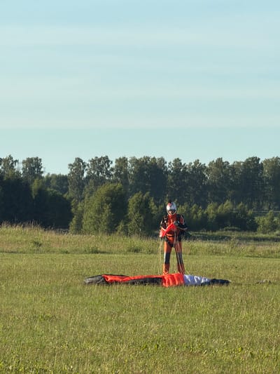 In a grassy field, a person in a bright orange and black skydiver suit stands holding red and white parachute lines. A folded parachute lies on the grass in front of them. The field is bordered by a dense line of trees under a pale blue sky.