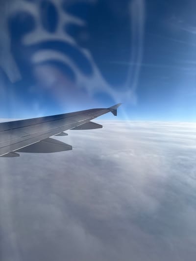 An aerial view from an aircraft window depicts the upper surface and leading edge of a metallic left-wing, featuring a distinct winglet and visible flap/slat structures. The aircraft is flying above a continuous, dense layer of white and light grey clouds that extends across the lower portion of the image, obscuring any ground features. The upper section of the scene displays a clear, vibrant blue sky with significant sunlight and lens flare effects originating from the upper right. Faint, blurred reflections of internal cabin elements are visible on the window's surface in the upper left quadrant. The flight is reportedly positioned over the city of Pozarevac, Serbia. No individuals or specific actions are discernible within the frame.