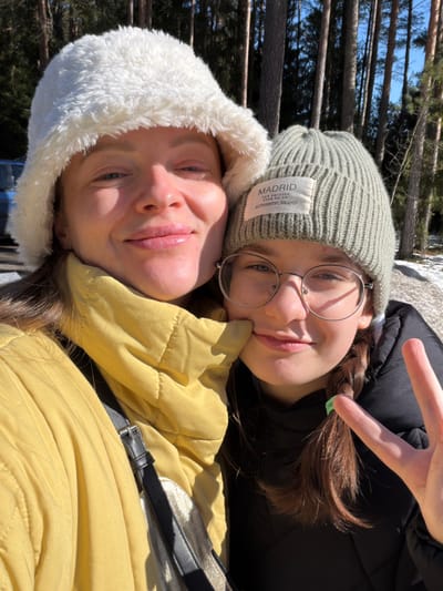 A close-up outdoor portrait features two females in a wooded area of Ракаўскі сельскі Савет, Belarus, under bright sunlight with patches of snow on the ground. The individual on the left, an adult female with light skin, wears a white faux-fur bucket hat and a yellow quilted jacket, smiling directly at the camera. The individual on the right, a younger female, possibly a teenager, with light skin and dark brown braided hair, wears round metal-rimmed glasses, a light olive green ribbed beanie with a "MADRID" label, and a black quilted jacket. She smiles and makes a peace sign gesture with her right hand, while her left hand gently touches the adult's lower left cheek. Tall pine trees and a clear blue sky are visible in the background, along with a partially obscured light-colored vehicle on the far left.