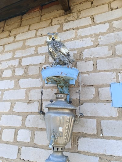 The image shows a metal samovar with an owl perched on top of it, mounted on a brick wall. The brick wall is made of light-colored bricks. The samovar is silver, with a blue painted metal base. The owl is grey and white with yellow eyes. The location is Уральский, Russia.