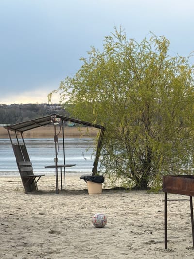 The image captures a riverside scene in Olginskaya, Russia. A light-colored sandy beach occupies the foreground, with a soccer ball resting on it. The beach transitions into the water's edge where a simple, metal-framed structure with a partially corrugated roof stands, acting as a shelter or picnic area. A small wooden table is visible inside the structure. The river extends into the midground. Beyond the river, there is a shoreline with visible vegetation and buildings, suggesting a residential area. A large tree with light green foliage dominates the upper portion of the image. To the right, there is a barbecue grill. The sky is partially cloudy and blue.