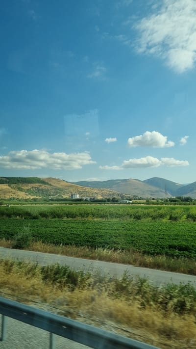 The image depicts a rural landscape under a bright, partly cloudy sky. In the foreground, a metallic guardrail is visible at the bottom, suggesting the photo was taken from a moving vehicle or elevated position. Beyond the guardrail, dry, yellowish grass lines a gray asphalt road that curves through the scene.
The middle ground is dominated by a vibrant green field, likely agricultural, stretching across the frame. Behind this field, rolling hills rise, covered in a mix of dry, golden vegetation and patches of green trees. In the distance, a cluster of low-rise buildings, possibly industrial or agricultural structures, are situated on the lower slopes of the hills. The background is filled with undulating mountains under a clear blue sky with scattered white clouds.
The scene conveys a sense of a sunny day in a rural area, possibly during late summer or early autumn given the dry grass and golden hills. The presence of the road and guardrail implies it is a man-made route through the natural landscape. The image is a static, wide shot capturing the expanse of the countryside. No people or animals are visible in the frame. The location is described as Krinitsa, Greece, which is known for its scenic countryside.