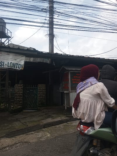 An outdoor street scene in Chori, Nigeria, is depicted under an overcast sky. In the foreground, the rear of a green motorcycle is visible, carrying two individuals. The passenger, seated closest to the camera, wears a dark red hijab, a light-colored, ruffled long-sleeved top, and blue jeans, with a patterned light purple scarf draped over her shoulders. A second person, likely the driver, is partially visible, wearing a dark hooded garment.
The background features several structures. To the left, a building with a white banner displaying the partial text "N_SI - LONTON" (suggesting "Nasi Lontong") stands beneath a corrugated metal roof. Above this building, a black water tank rests on a metal stand. Adjacent to this, a stone wall segment with a dark green metal gate is present. To the right of the gate, a white-framed food stall with red lettering advertises "SEDIA", "SATE AYAM", "SATE KAMBING", and "SOP KAKI" (chicken satay, lamb satay, and leg soup). Behind and above the buildings, a tall, grey utility pole is positioned centrally, from which numerous black electrical and communication wires extend horizontally across the frame against the grey sky. The ground consists of a rough asphalt road in the immediate foreground, transitioning to a cracked concrete path leading to the buildings, with a dark, elongated metal object lying on the concrete.