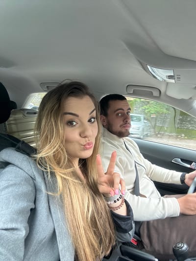 Two individuals are inside a vehicle. The location is suggested to be Stara Kresna, Bulgaria, based on the surrounding environment seen through the car window. A woman is in the foreground, partially visible with light brown and blonde hair, wearing a gray hooded sweatshirt. She is making a peace sign with her hand and has a duck-lipped expression. Behind her, a man is partially visible, with dark hair and a beard. He is wearing a light-colored jacket with a black shoulder bag. The interior of the vehicle is visible, with light-colored ceiling and door panels. The steering wheel and dashboard are in the background. The scene appears to be captured from a phone camera.