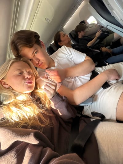 Inside an airplane cabin, a group of people are seated. The camera focuses on a young woman with blonde hair, reclined with her eyes closed. Beside her is a young man with brown hair, gazing at the camera. They are seated closely, holding hands. The woman wears a brown sweatshirt and the man wears a white t-shirt. Other passengers are visible in the background, some using their phones. The airplane window is to the left.