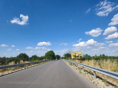 The image depicts a road in Krushovitsa, Bulgaria, on a clear, sunny day with a bright blue sky dotted with white, puffy clouds. The road is asphalt with a single orange line marking the center. Guardrails line both sides of the road, with vegetation, including trees and dry grass, visible beyond them. In the distance, two cars are driving on the road. A yellow directional sign is positioned on the right side of the road, with text in Bulgarian indicating directions to Pleven and Yasen. The sign also shows "3.5T" indicating a weight limit, and is partially covered in graffiti. The overall scene suggests a rural or semi-rural setting.