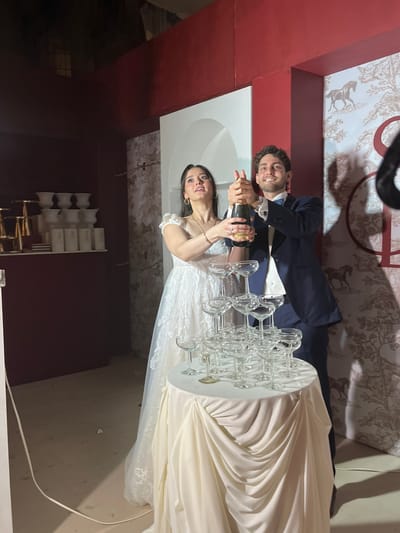 A wedding scene is captured indoors in what appears to be Orizaba, Mexico, judging from possible architectural and decorative styles. A couple, dressed in wedding attire, is positioned in front of a tower of clear wine glasses placed on a round, white table. The man, wearing a dark suit, is holding a champagne bottle and assisting a woman in a white wedding dress to open the bottle, presumably to pour into the glasses. The backdrop includes a textured wallpaper or wall design in muted tones. The location appears to be a reception venue with dark walls and architectural details.