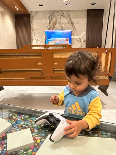 The image displays a young child interacting with a video game controller in an indoor setting. The image is oriented vertically (rotated 90 degrees clockwise from its natural upright position).
The central focus is a young child, likely a toddler or preschooler, with dark, somewhat messy hair. The child is wearing a two-toned long-sleeved shirt: the main body is light blue, and the sleeves and collar are yellow. The brand name "adidas" is prominently displayed in yellow text on the blue portion of the shirt. The child's face is looking downwards, with eyes appearing to be closed or cast down intently. Their left hand is resting on a white PlayStation 5 DualSense controller, which is placed on a clear glass table. The child's right hand is slightly clenched.
The glass table beneath the controller contains a decorative layer of numerous small, polished, and colorful stones or marbles. These stones are mostly iridescent or translucent, with dominant hues of blue, green, and pink, creating a vibrant base beneath the transparent surface.
In the background, behind the child and the table, there is a piece of light brown wooden furniture, possibly a cabinet or part of a bed frame, featuring vertical slat details. Beyond this furniture, a wall with a prominent white and gray marble pattern is visible. Mounted on this marble wall is a flat-screen television, which is actively displaying content. The screen shows bright blue and purplish colors, suggesting a video game or animated program.
Below the marble wall, there's a horizontal band of dark brown paneling, possibly made of wood or a similar material with a textured, slatted appearance. To the far left side of the image, a white wall is visible, on which a white device resembling a small security camera or sensor is mounted. Further down on this white wall, two small, circular, dark fixtures (possibly light sources or outlets) are visible. The floor appears to be a light-colored, possibly tiled, surface.
The lighting suggests an indoor setting, likely daylight or bright artificial light. The overall scene conveys a moment of focused engagement as the child interacts with the gaming controller.