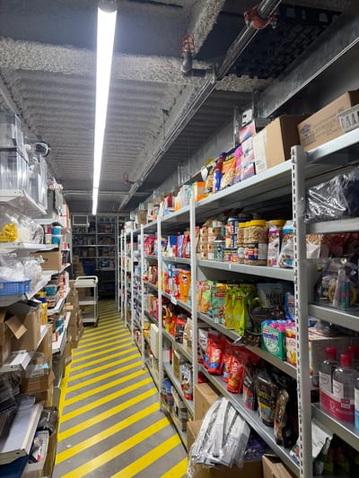 This image captures an interior view of what appears to be a warehouse or a large storage area, possibly a distribution center or a large retail store backroom, in Longueuil, Canada. The perspective is looking down a long aisle filled with shelves stacked high with various packaged goods.

The primary subjects are the extensive metal shelving units packed with a wide assortment of products, predominantly snack foods like nuts, chips, and candies, alongside some personal care or over-the-counter items. The shelves are tightly packed, indicating high-density storage. Below the shelves, on the floor, are numerous cardboard boxes and clear plastic containers, suggesting items are either being prepared for shipment, organized, or recently received. The floor features distinctive diagonal yellow and gray stripes, commonly used for demarcation or safety in industrial settings.

Notable details include a bright, elongated fluorescent light fixture mounted on the left wall, illuminating the aisle. The walls have a textured, possibly sound-dampening or insulation material, and industrial pipes run along the upper section of the wall and ceiling. No people are visible, and no specific activity is taking place, but the scene implies ongoing logistics and inventory management. The indoor setting means time of day and weather are not discernible.

Visible text on packaging and boxes includes product names and brands such as "PLANTE," "HARVEST SNAP," "mentos PUREFRESH Extra," "PRANA fuji," "CRISPY FENELS," "Stover," "ERMA" (likely Derma or a similar brand, seen on red-capped bottles), and "Carte5-". Other legible text includes "Gout de BBQ Piquant Amandes 10943" and "ZESTY BBQ VALUE PK O" on cardboard boxes, along with various numbers like "30," "37,403," and "12,495." French language on some packaging is consistent with the Longueuil, Canada location context.
