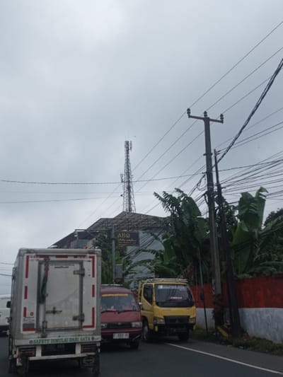 A street scene under a cloudy sky shows a white truck in the foreground, followed by a red car and a yellow truck. A tall cell tower rises behind a building with an "ABQ" sign. Power lines and a utility pole are visible. A wall painted with red and white stripes runs along the side of the road, adjacent to banana trees.
