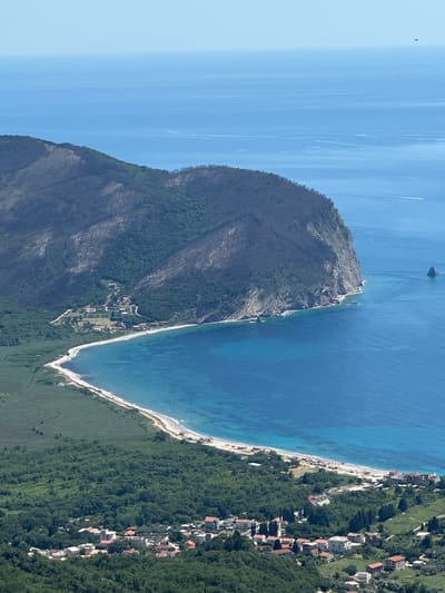 An aerial shot displays a coastline and mountainous terrain. The ocean is a gradient of blues, transitioning from lighter near the horizon to darker near the shore. A prominent headland extends into the sea, covered in vegetation and scorched patches. A narrow beach separates the water from the land. Buildings dot the coastline, interspersed with green trees. A structure in the water, perhaps a boat, is visible in the distance. The overall impression is one of a coastal landscape. This is Novoselje, Montenegro.