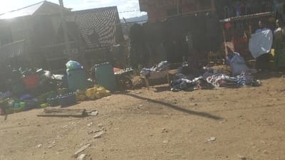 The image shows a street scene in Kayna, Democratic Republic of the Congo. It depicts a dirt road in front of several buildings. The ground is covered in various items, including plastic containers, clothing, and fabric. Two large blue plastic containers are on the left side of the road, while a wooden structure with a white cloth draped over it sits on the right side. A person is standing behind the structure, wearing a white shirt and black pants. They are holding a white cloth in their hands. There are other people in the background but they are not visible.  A large black sheet hangs on the wall of a building in the background.  The image appears to be taken in daylight.