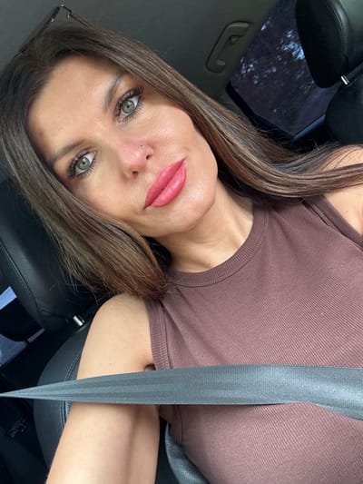 A woman with brown hair and light green eyes is seen from the chest up, sitting in the driver's seat of a car. She is wearing a brown ribbed tank top. A grey seatbelt is across her chest. Her left arm is visible below the seatbelt. The interior of the car, including a black leather seat and part of the roof, is visible. The background shows a dark, blurry image of trees and the sky through the car's sunroof.